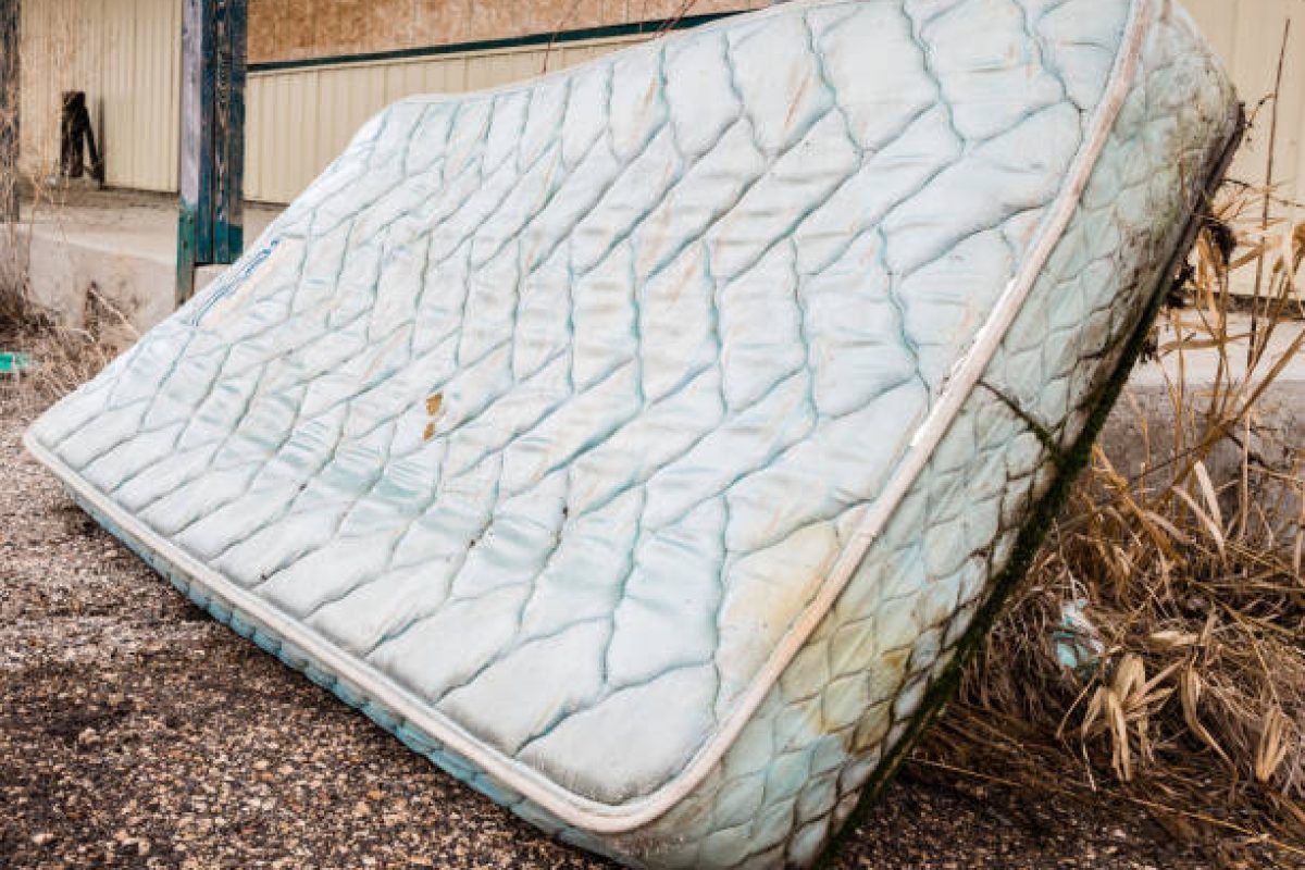 horizontal image of an old worn out water logged mattress leaning against a railing in the summer time